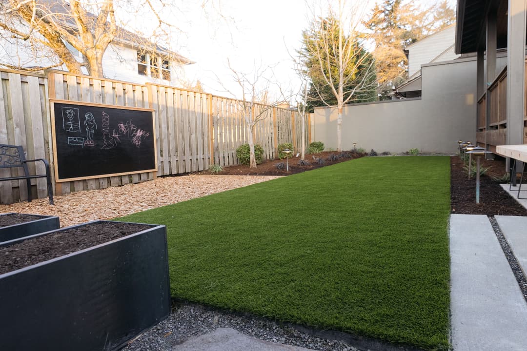 Lush green artificial grass in backyard with chalkboard, garden beds, and decorative plants.