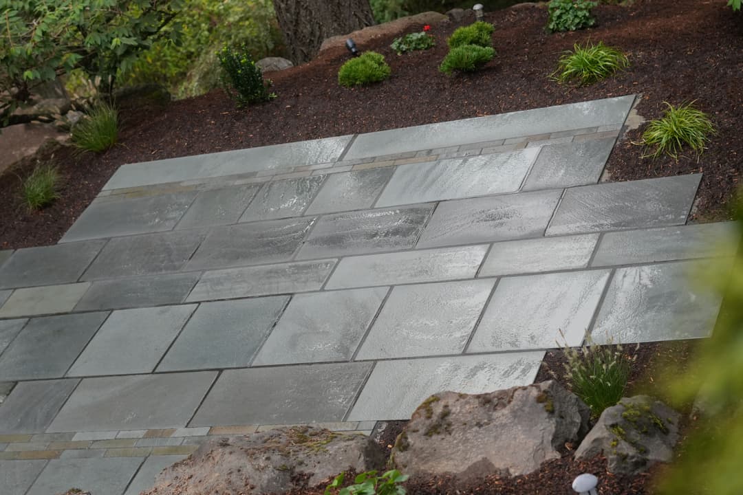 Wet natural stone patio with intricate patterns, surrounded by decorative plants and mulch.