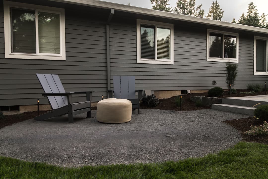 Outdoor seating area with two gray chairs and a round ottoman beside a modern house.