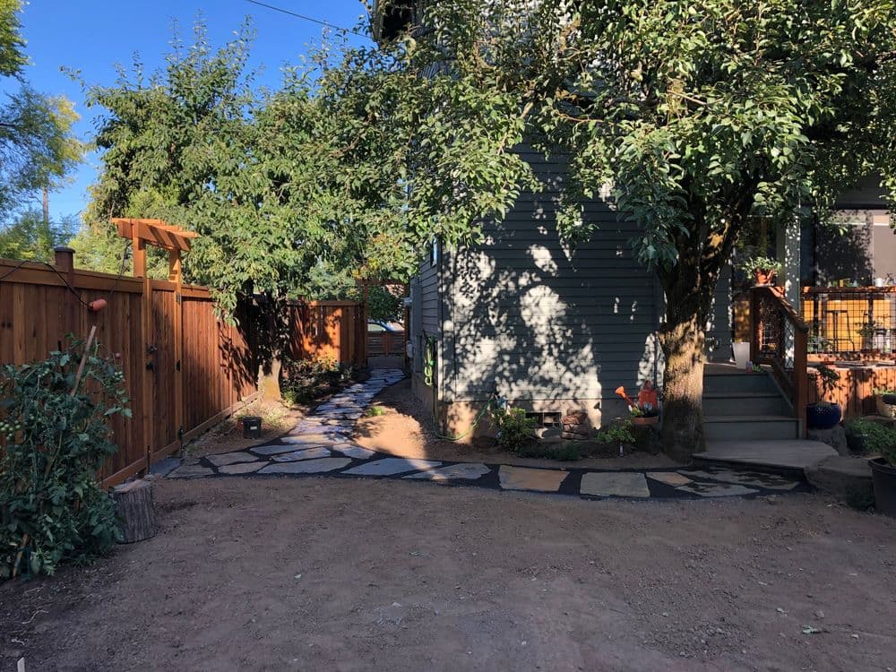 Garden pathway beside a home, featuring stone pavers, trees, and wooden fence.