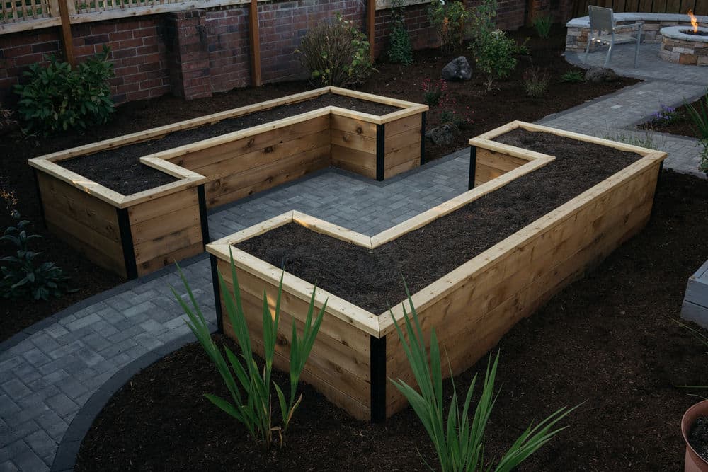 L-shaped wooden raised garden beds in a landscaped yard with stone path and fire pit.