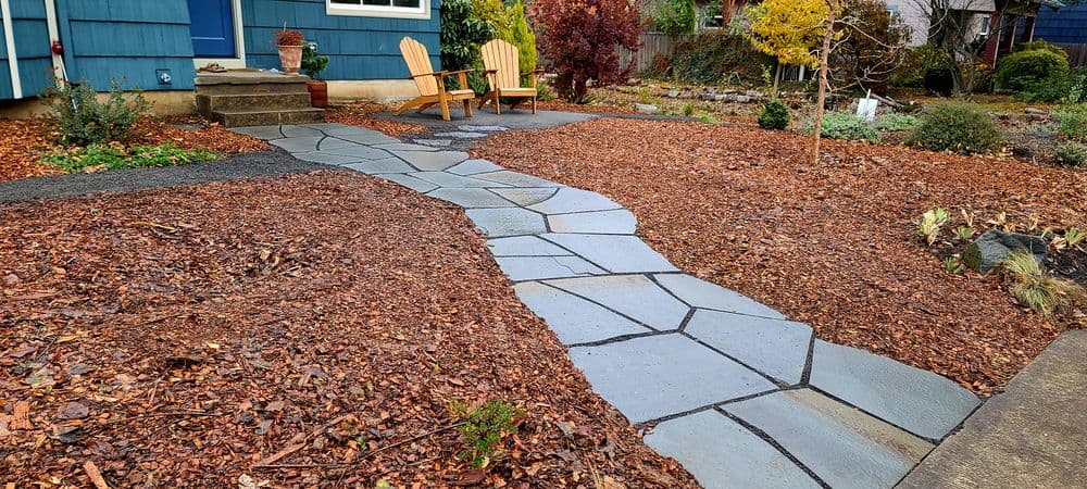 Stone pathway winding through mulch landscape with wooden chairs in a garden setting.