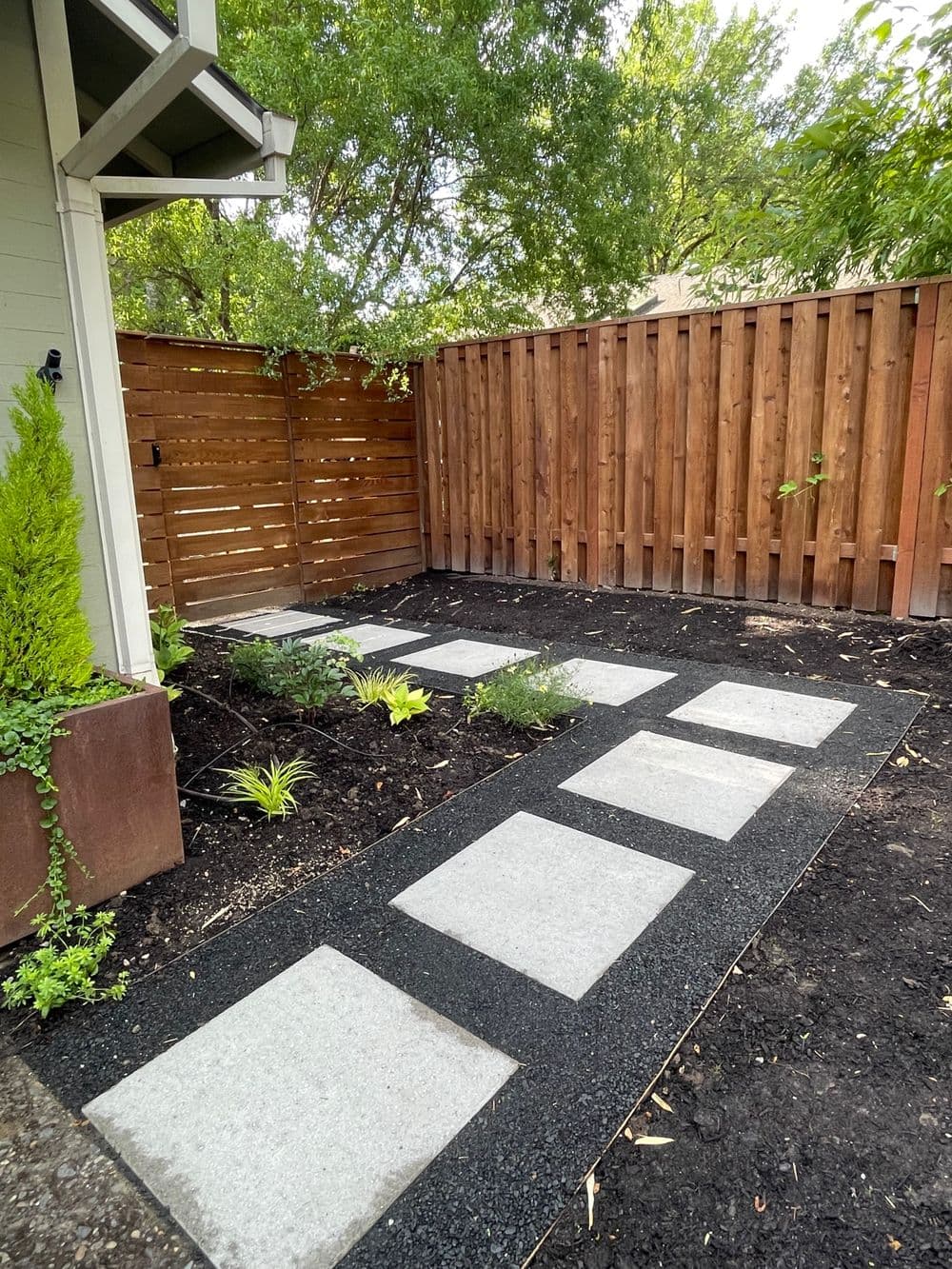 Modern garden pathway with concrete stepping stones, bordered by greenery and wooden fence.