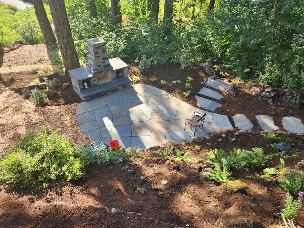 Stone patio with fireplace, surrounded by greenery and garden beds in a wooded area.