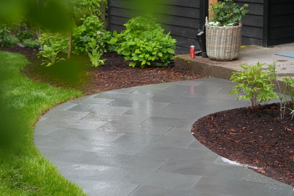 Curved gray stone pathway in a garden with lush greenery and freshly mulched flower beds.