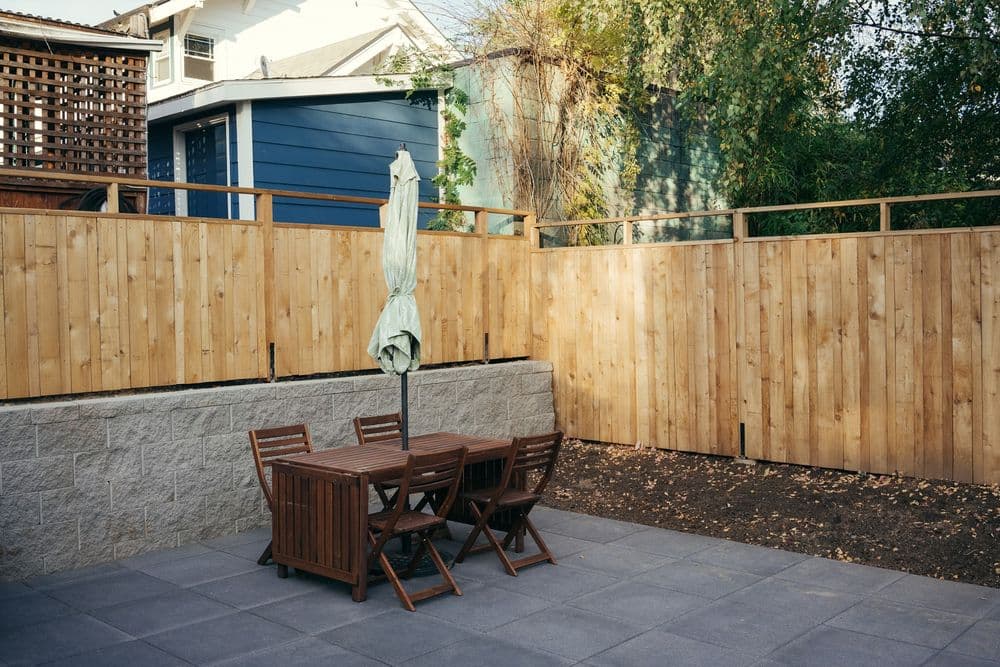 Outdoor patio with wooden dining set and umbrella, surrounded by wooden fencing and greenery.