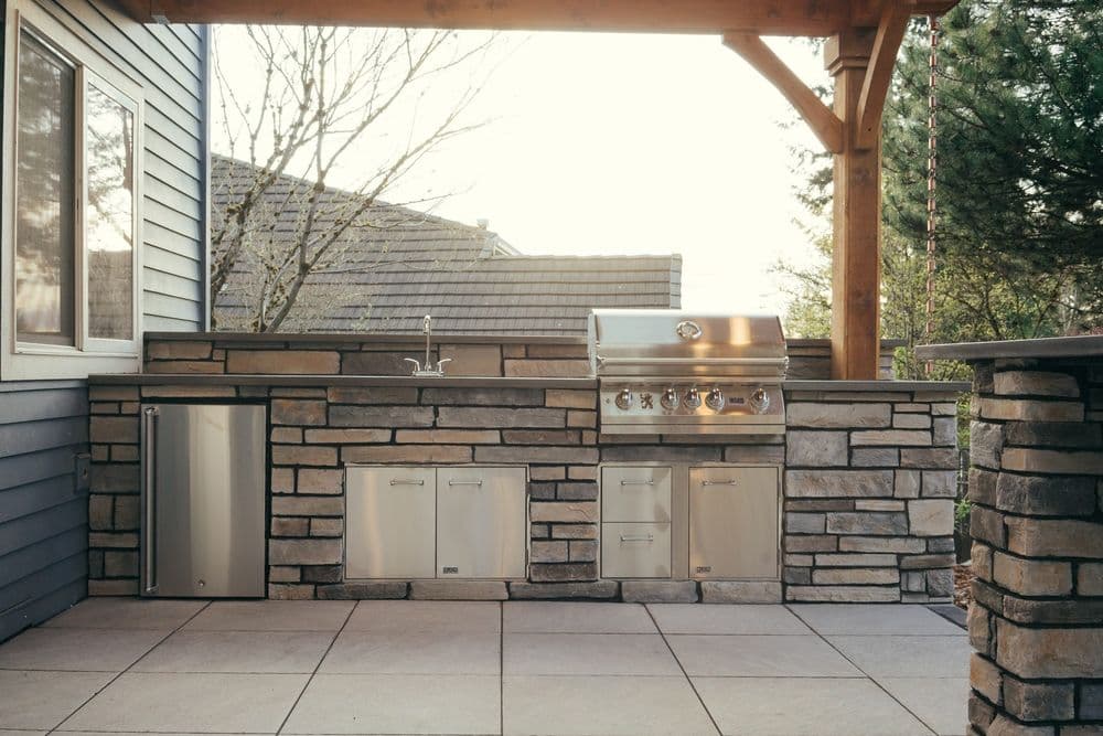 Outdoor kitchen featuring a stone grill station, sink, and storage under a wooden pergola.