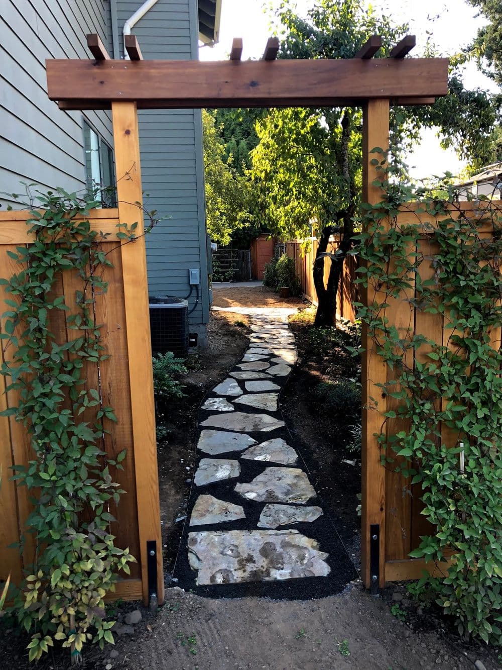 Garden pathway with stone tiles leading through wooden gate to lush greenery and trees.