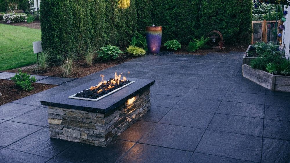 Modern outdoor patio featuring a stone fire pit with flames, surrounded by greenery.
