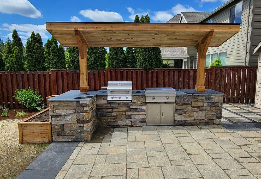 Outdoor kitchen with stone grill station, wooden pergola, and paved patio in a backyard setting.