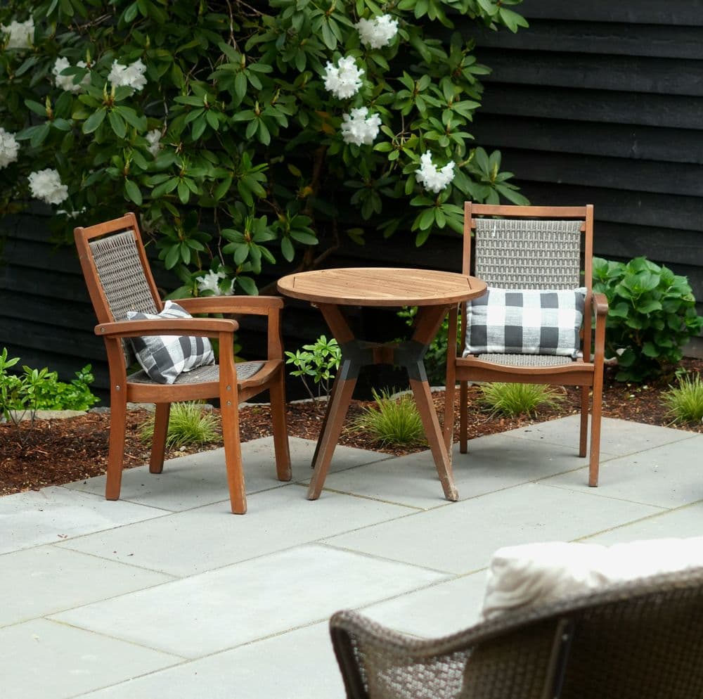 Outdoor patio scene with wooden chairs and a round table surrounded by greenery and flowers.