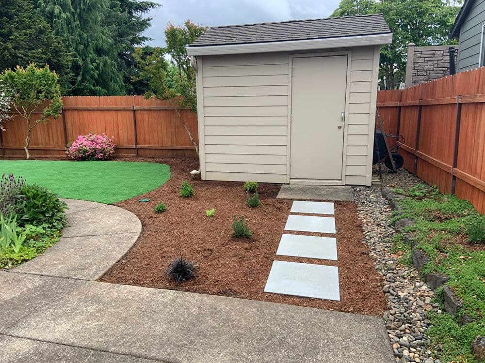 Shed in landscaped yard with grass, mulch, and stepping stones surrounded by wooden fence.