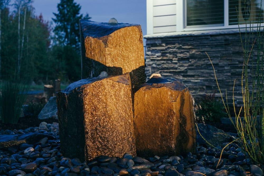 Illuminated stone fountain near a house, surrounded by pebbles and plants at dusk.