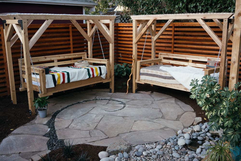 Stylish outdoor swinging beds on a stone patio surrounded by wooden fencing and plants.