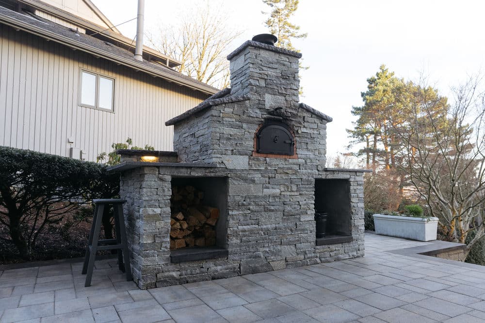 Stone outdoor fireplace with wood storage and chimney in backyard patio setting.