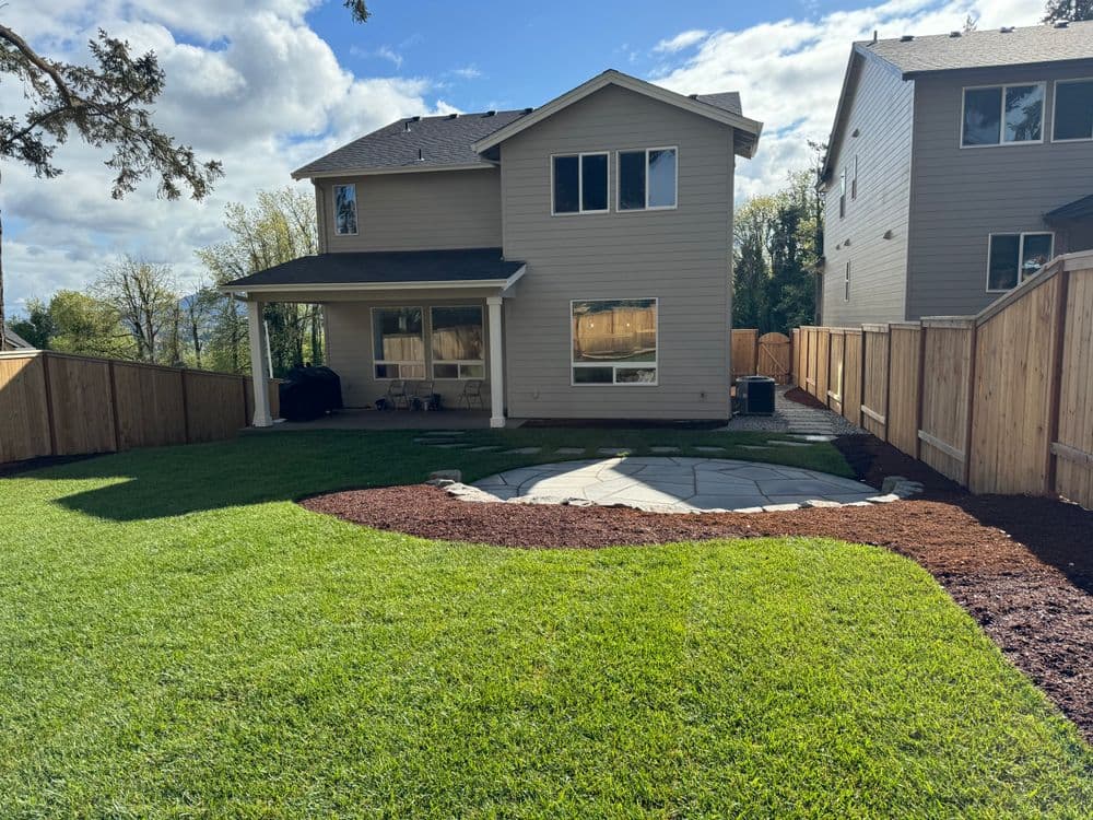 Backyard view of a modern home with green grass, stone patio, and wooden fence.