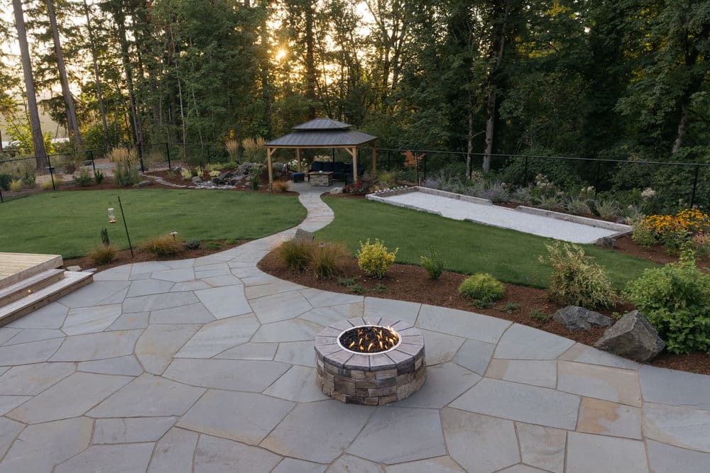 Beautiful backyard landscape featuring a stone fire pit, gazebo, and landscaped garden at sunset.