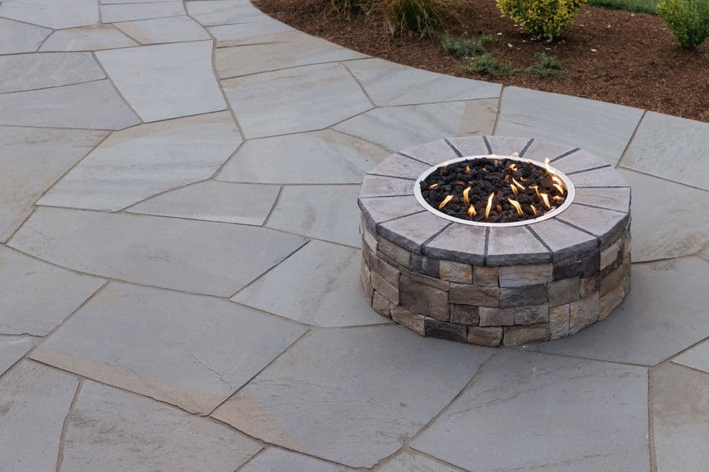 Modern stone fire pit with flames on a tiled patio, surrounded by lush landscaping.