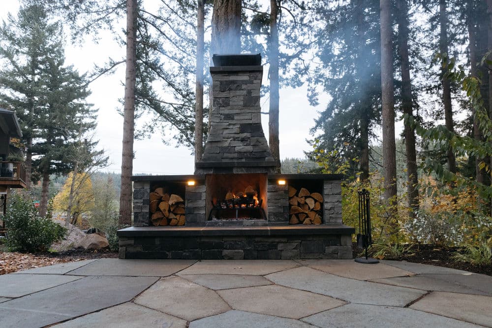 Outdoor stone fireplace with smoke, surrounded by trees and stacked firewood on a patio.