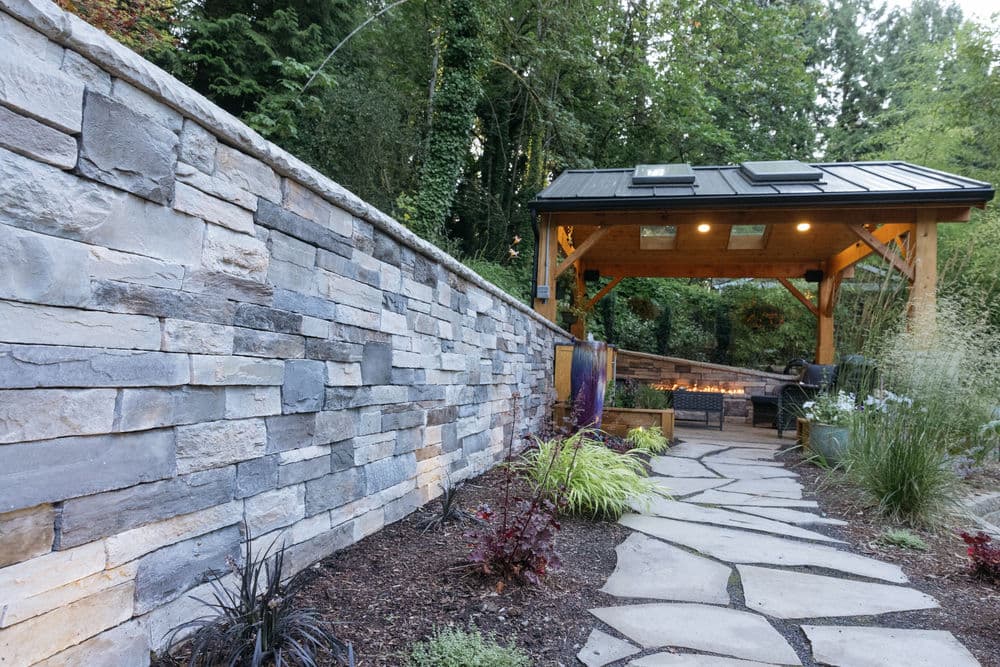 Stone garden path leading to a wooden gazebo with a fire pit and lush greenery.