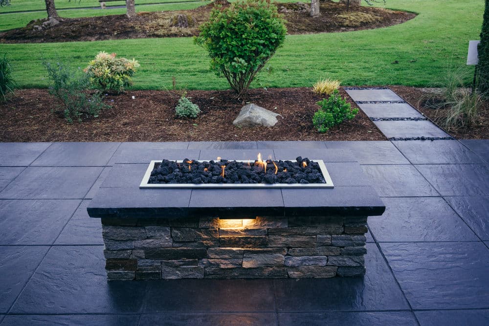 Outdoor fire pit with black lava rocks and modern stone base, surrounded by a landscaped yard.